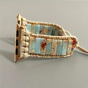 Könnte beinhalten: Ein Uhrenarmband mit rechteckigen, hellblauen und rotbraunen Steinperlen, die mit cremefarbener Kordel und goldfarbenen Metallketten verflochten sind. Das Band enthält ein goldfarbenes Metallverbindungsstück zur Befestigung an einer Uhr.