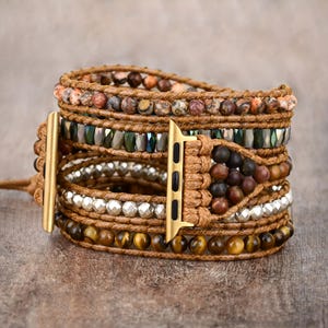 Könnte beinhalten: Mehrschichtiges Wickelarmband für eine Apple Watch. Es hat braune, grüne und silberne Natursteinperlen, umwickelt mit braunem Leder. Der Uhrenadapter ist goldfarben. Der Umfang beträgt ca. 18 cm.