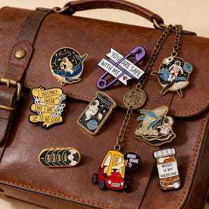 May include: A collection of enamel pins with various designs, including an astronaut, a whale, a safety pin with text, and a car. The pins are in various colors, including black, blue, yellow, and red. Some pins have text such as "You are safe with me" and "Music is my medicine."