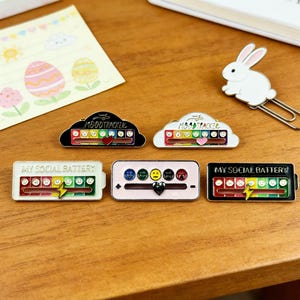 Op de afbeelding: Een verzameling emaille pins met stemming- en sociale batterij-indicatoren. De pins hebben verschillende ontwerpen, waaronder wolkvormen met de tekst "Moodtracker" en batterijvormige pins met de tekst "My Social Battery". Elke pin heeft een schaal met emoji-gezichten.