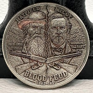 May include: A silver-toned commemorative coin featuring relief portraits of two men, labeled Hatfield and McCoy. Below them are two crossed rifles and the text BLOOD FEUD 1863-1891. The names Devil Anse Hatfield and Ole Ran'l McCoy are inscribed.