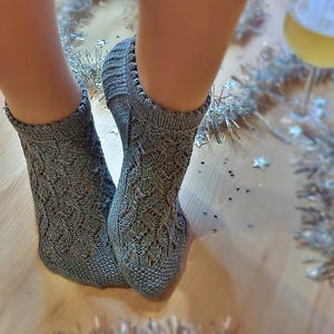 Könnte beinhalten: Nahaufnahme von grauen Strickstrümpfen mit einem detaillierten Lochmuster. Die Socken werden an Füßen getragen, darunter ist ein Holzboden zu sehen. Die Socken haben einen gerippten Bund und ein dekoratives Design. Die Szene ist festlich, mit silbernem Lametta und sternförmigem Konfetti.
