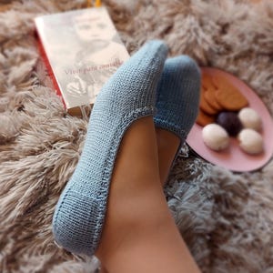 Puede incluir: Calcetines de zapatilla de punto azul claro. Los calcetines se usan con un libro y un plato de galletas y caramelos sobre una alfombra suave de color marrón claro. El libro tiene el título "Vivir para contarlo".