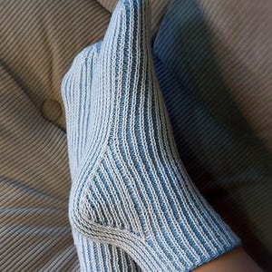 Puede incluir: Un par de calcetines tejidos con un patrón de canalé vertical en rayas alternas de color azul claro y blanco. Los calcetines tienen una construcción de talón definida y una silueta ajustada que se extiende desde el tobillo hasta la punta.