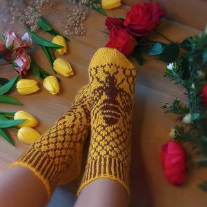 Könnte beinhalten: Ein Paar gelb-braune Stricksocken mit einem Bienenmuster. Die Socken haben ein geometrisches Muster und ein detailliertes Bienenmotiv auf der Vorderseite. Die Abbildung zeigt Blumen, darunter gelbe Tulpen und rote Rosen.