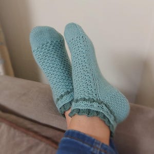 May include: A pair of light blue knitted socks with a textured pattern and a ruffled edge. The socks are worn on a person's feet, with the legs and feet visible. The socks are a soft, muted blue color.