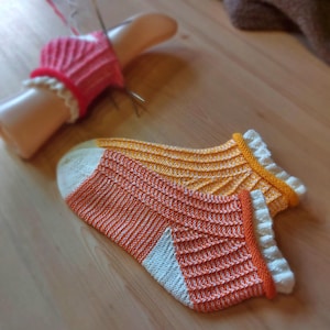 May include: A pair of hand-knitted orange and white socks with a ribbed design and white trim. A pink knitted wrist warmer with white trim is on a mannequin hand. The socks are displayed on a wooden surface.
