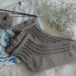 May include: A pair of beige knit socks with a ruffled cuff and textured stitch pattern, placed next to a cream-colored t-shirt featuring a blue moon and cloud graphic, a ball of beige yarn, and two knitting needles.