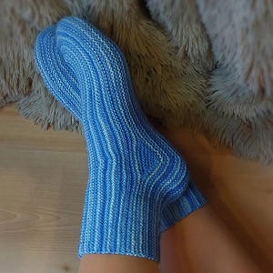 Könnte beinhalten: Ein Paar handgestrickte Socken mit einem melierten Muster aus hellblauem, mittelblauem und weißem Garn. Die Socken haben eine gerippte Textur, die vertikal entlang des Fußes und Knöchels verläuft und in einer verstärkten Ferse und Spitze endet.