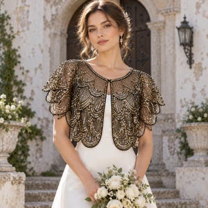 May include: A black bolero jacket featuring intricate gold sequin embroidery in a scalloped, layered pattern. The garment has short, tiered sleeves and a rounded neckline that fastens at the center, worn over a plain white dress.