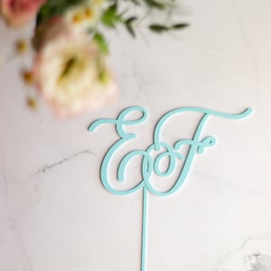 May include: A pale blue cake topper with the intertwined letters "E" and "F" in a cursive font. The topper has a single, thin stem for insertion into a cake. Soft focus flowers are in the background.