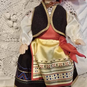 Könnte beinhalten: Eine Vintage-Puppe in traditioneller Volkskleidung. Die Puppe hat ein bemaltes Gesicht, schwarzes Haar und trägt ein rotes Kopftuch. Ihr Outfit umfasst eine schwarze Weste, eine weiße Bluse und einen gemusterten Rock mit einem goldenen und roten Schärpenband.