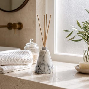 May include: A conical white marble reed diffuser with grey veining, featuring a small white neck and four thin, light-colored wooden reeds extending from the top. The object has a smooth, polished stone texture and a wide, stable base.