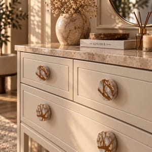 May include: A light beige wooden dresser featuring four round drawer knobs with a white and brown marble pattern. The dresser has a matching light beige marble countertop. On top sits a round marble vase, a book titled The Kinfolk Home, and a reed diffuser.