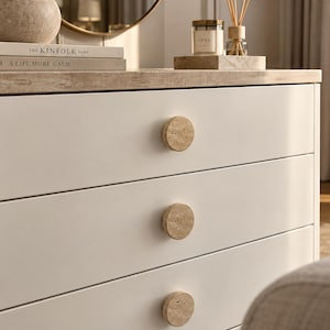 May include: A white three-drawer dresser featuring circular, porous beige stone knobs. The top surface is a light tan stone, and the drawers are stacked vertically. The books titled The Kinfolk Home and A Life More Calm are visible.