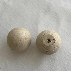 Op de afbeelding: Twee beige, bolvormige kastknoppen met een textuur, steenachtige uitstraling. Eén knop heeft een voorgeboord gat voor montage. De knoppen zijn waarschijnlijk voor meubels, wat een natuurlijke, rustieke esthetiek toevoegt.