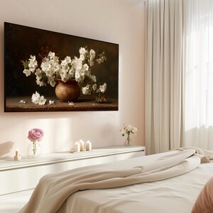 Puede incluir: Una pintura enmarcada representa una naturaleza muerta de flores blancas en un jarr&oacute;n marr&oacute;n. La obra de arte est&aacute; montada en una pared rosa claro sobre una c&oacute;moda blanca. Una sola rosa rosa en un jarr&oacute;n de cristal est&aacute; sobre la c&oacute;moda.