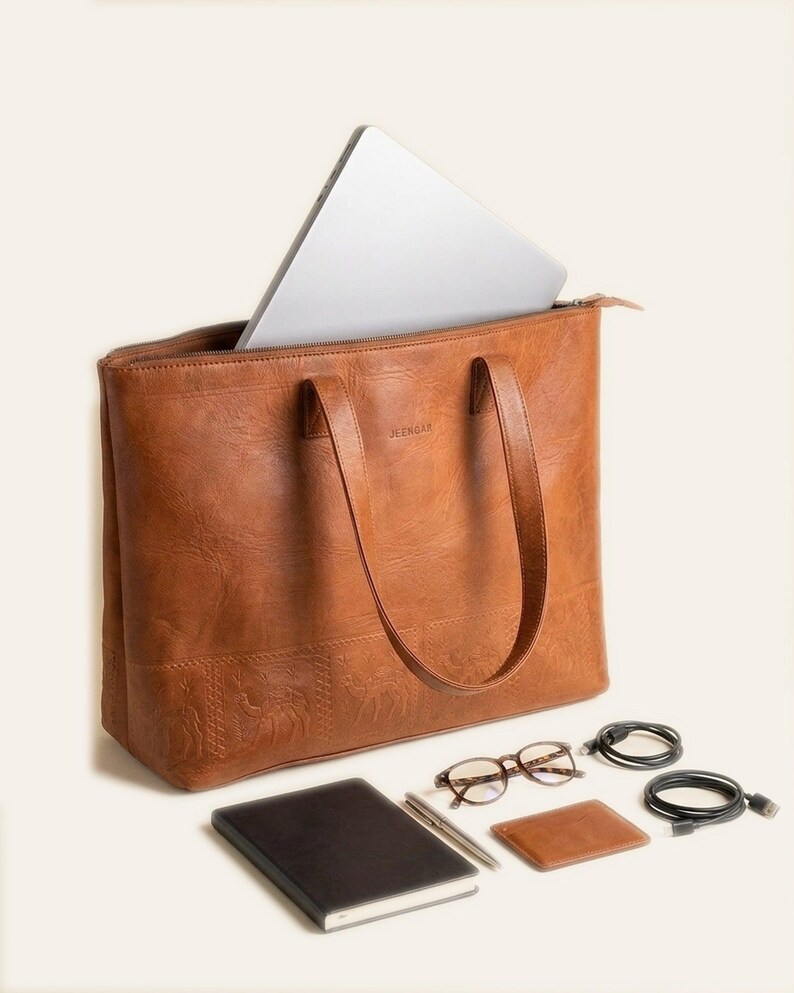 A 16-inch laptop sliding into The Abhaneri Tote, with a notebook, pen, glasses, cardholder and cables laid alongside