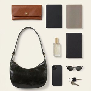 The Ranisar Shoulder Bag in dark olive, shown with a clutch, e-reader, paperback, perfume bottle, phone, sunglasses and keys to show what it holds (AI-generated)