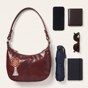 The Kashida Shoulder Bag, Padmini motif in Cherry, shown with a smartphone, a wallet, sunglasses, a compact umbrella and a pocket notebook to show what it holds (AI-generated)