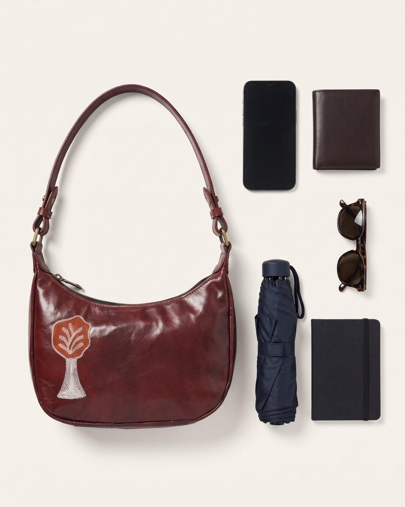 The Kashida Shoulder Bag, Padmini motif in Cherry, shown with a smartphone, a wallet, sunglasses, a compact umbrella and a pocket notebook to show what it holds (AI-generated)