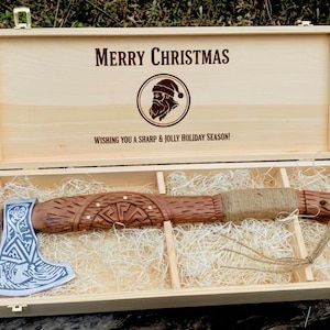 May include: Wooden box with "Merry Christmas" text and a Santa Claus graphic. Inside, a decorative axe with a silver blade and carved wooden handle rests on straw. The axe is a holiday gift.