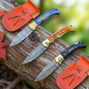 Puede incluir: Tres navajas plegables personalizadas con hojas de acero de Damasco y mangos de madera o de colores. Los mangos están grabados con nombres y el texto "ADVENTURE IN 2024". Cada navaja tiene una funda de cuero. Las navajas se exhiben sobre una superficie de madera.