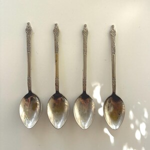 May include: Four silver-toned metal spoons arranged in a vertical row. Each spoon features a slender handle with an embossed figure at the top, a decorative geometric pattern below the figure, and a smooth, oval-shaped bowl with visible tarnishing.