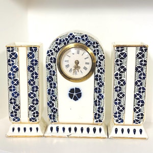 May include: A ceramic mantel clock and two matching rectangular vases featuring a white base with blue floral patterns and gold trim. The clock has a circular face with black Roman numerals and gold hands, centred within an arched frame.