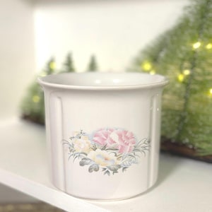May include: A white ceramic container with a floral design featuring pink and yellow flowers. The container has a slightly flared rim and vertical grooves on the sides. The floral arrangement is centered on the front.