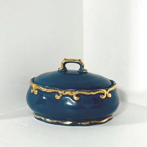 Puede incluir: Una caja de baratijas de cerámica azul oscuro con tapa y ribete dorado. La caja tiene forma redondeada con un pequeño asa en la parte superior. El ribete dorado decora los bordes de la tapa y la base.