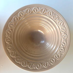 Może przedstawiać: Okrągła, jasnobeżowa miska ceramiczna z szerokim, płaskim brzegiem ozdobionym powtarzającym się tłoczonym wzorem kwiatowym. Wnętrze miski posiada szereg koncentrycznych kolistych rowków, które schodzą w kierunku środkowej podstawy.
