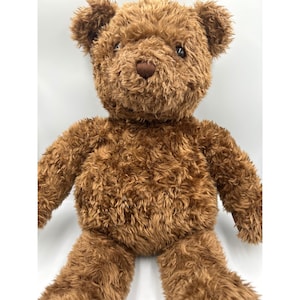 May include: A brown teddy bear with a soft, plush texture. The bear has a heart-shaped nose and small, dark eyes. Its fur is a warm, medium brown colour, and it has a classic teddy bear design.