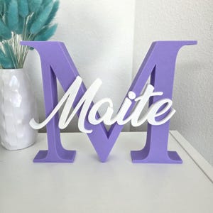 May include: A lavender-colored letter "M" with the name "Maite" in white script across the front. The decorative piece is on a white surface, with a white vase and blue decorative stems in the background.