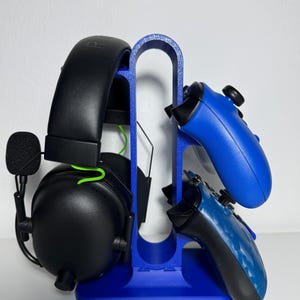 Pode incluir: Suporte azul para fone de ouvido para jogos com um fone de ouvido preto e dois controles de jogo azuis. O suporte é feito de plástico azul e tem um design curvo. O fone de ouvido tem um cabo verde.