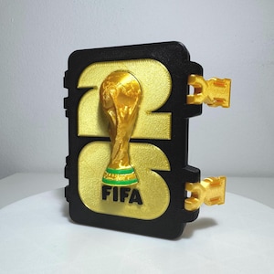 May include: A black rectangular plaque featuring the gold-colored number 26 with a gold FIFA World Cup trophy emblem integrated into the design. The trophy base has a green band, and the word FIFA is printed in black below it. Two gold side clips.