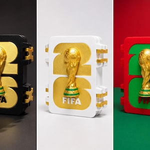 Pode incluir: Três estojos retangulares com o número 26 e um emblema dourado do troféu da Copa do Mundo da FIFA no centro. O primeiro estojo é preto com detalhes dourados, o segundo é branco com dourado e o terceiro é vermelho com detalhes verdes e troféu.