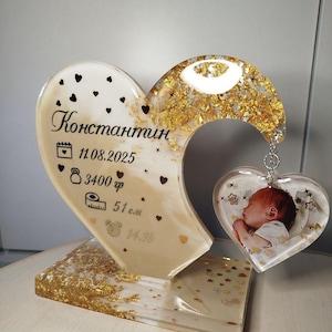 May include: A heart-shaped resin keepsake with gold leaf accents and a small heart-shaped photo pendant. The main heart has the name "Konstantin" and birth details. The base is rectangular with gold leaf.