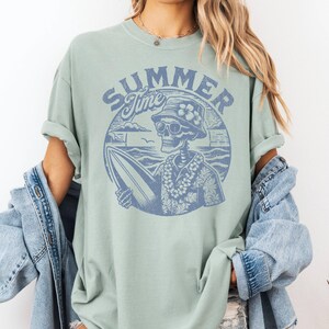 May include: A light green t-shirt with a blue graphic of a skeleton surfer holding a surfboard, wearing sunglasses, a floral lei, and a bucket hat. The words "SUMMER Time" are above the skeleton in a circular design.