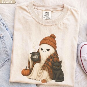 May include: An ivory t-shirt featuring a graphic of a ghost wearing an orange knit beanie and a plaid blanket, holding a black mug with the text NOPE. A small orange pumpkin and a black cat sit beside the ghost. The shirt has a crew neckline.