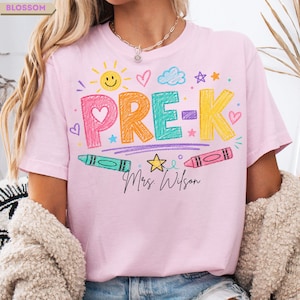 May include: A light pink t-shirt featuring the text PRE-K in large, multi-colored crayon-style letters. The design includes a yellow sun, blue cloud, stars, hearts, and two crayons. Below the text, the name Mrs. Wilson is printed in black script.
