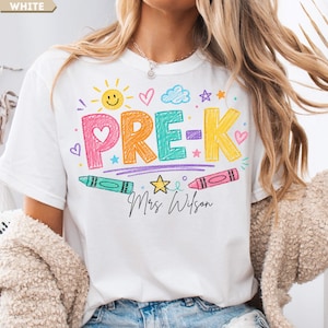 May include: A white t-shirt featuring a colorful graphic with the text PRE-K and Mrs. Wilson in black script. The design includes crayon-style lettering in pink, orange, teal, and yellow, surrounded by drawings of a sun, clouds, stars, and hearts.