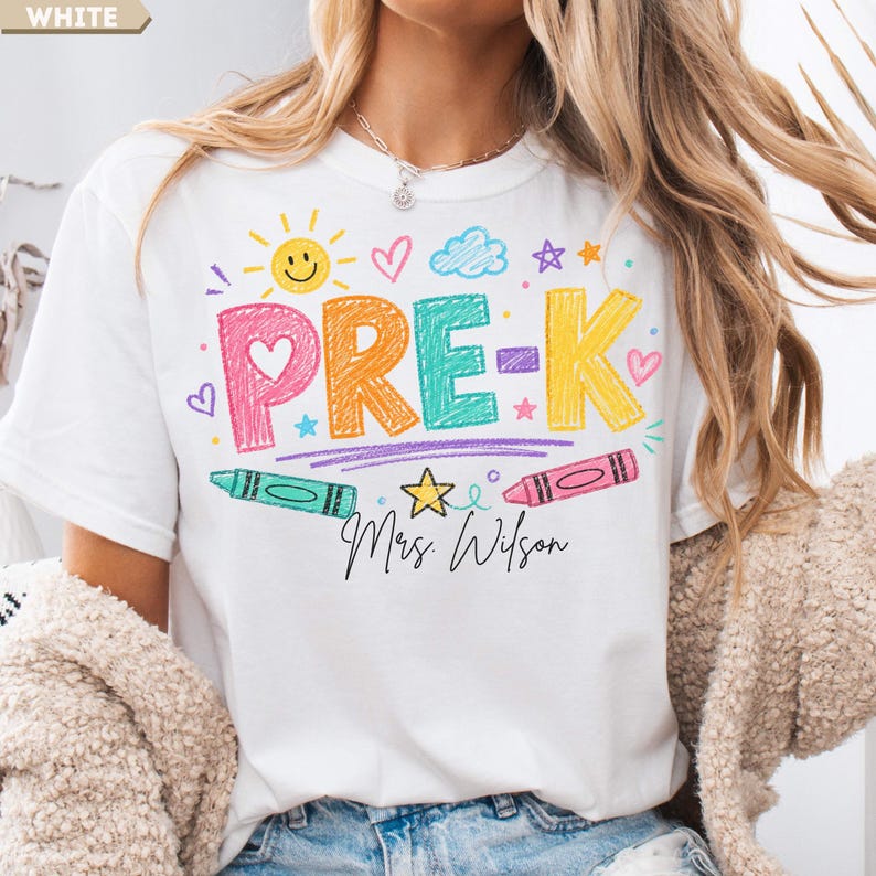 May include: A white t-shirt featuring a colorful graphic with the text PRE-K and Mrs. Wilson in black script. The design includes crayon-style lettering in pink, orange, teal, and yellow, surrounded by drawings of a sun, clouds, stars, and hearts.
