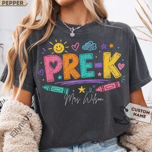 May include: A charcoal gray t-shirt featuring the colorful text PRE-K in a crayon-like font. The design includes a yellow sun, hearts, stars, clouds, and two crayons. Below the text, the name Mrs. Wilson is written in a white script font.