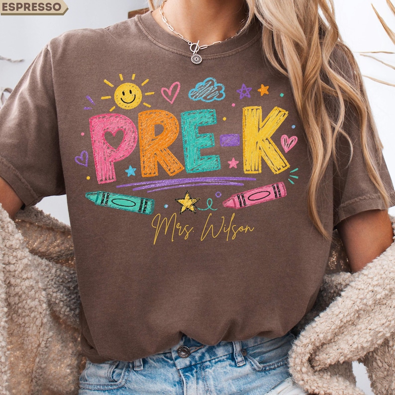 May include: A brown short-sleeve t-shirt featuring the text PRE-K in colorful, textured letters. The design includes a yellow sun, hearts, stars, clouds, and two crayons. Below the text, the name Mrs. Wilson is written in yellow cursive script.