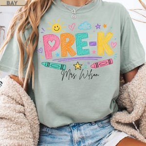 May include: A sage green t-shirt featuring a colorful graphic with the text PRE-K in a crayon-like texture. The design includes a yellow sun, hearts, stars, clouds, and two crayons. Below the graphic, the name Mrs. Wilson is printed in black script.