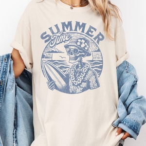 May include: Cream-colored t-shirt featuring a blue graphic of a skeleton surfer with sunglasses, a floral hat, and a lei, holding a surfboard. The words "SUMMER Time" are arched above the skeleton in a circular design. The shirt has short sleeves and a relaxed fit.