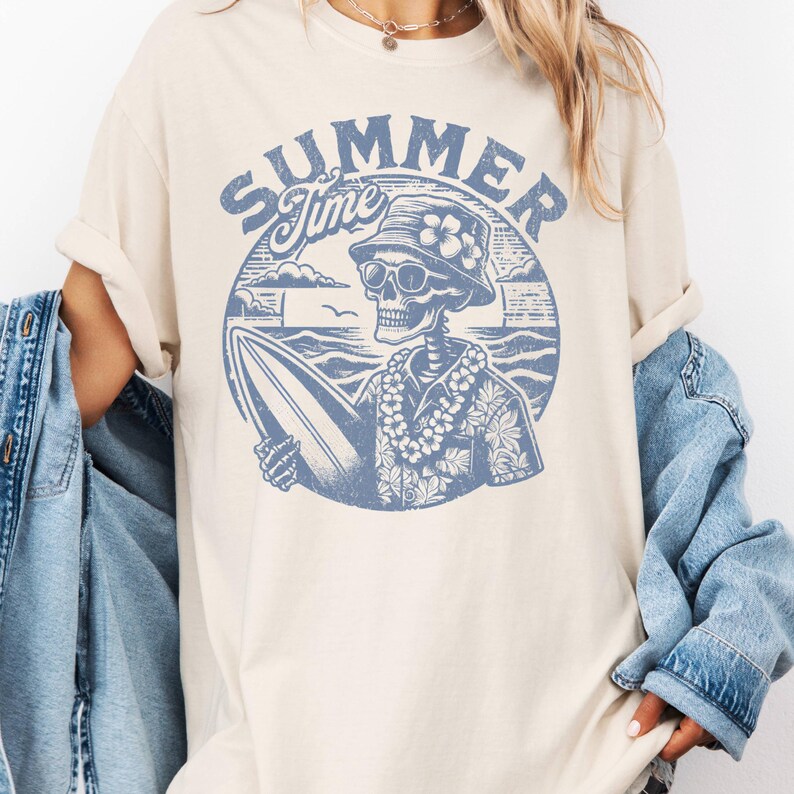 May include: Cream-colored t-shirt featuring a blue graphic of a skeleton surfer with sunglasses, a floral hat, and a lei, holding a surfboard. The words "SUMMER Time" are arched above the skeleton in a circular design. The shirt has short sleeves and a relaxed fit.