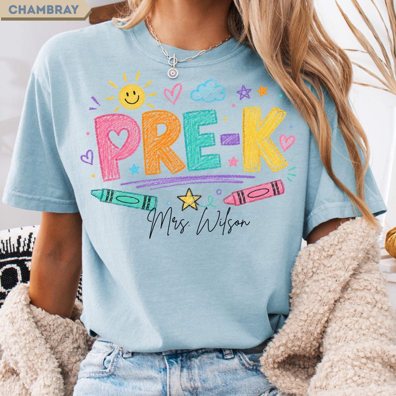 May include: A light blue t-shirt featuring the text PRE-K in colorful, crayon-textured letters. The design includes a yellow sun, a blue cloud, stars, hearts, and two crayons. Below the text, the name Mrs. Wilson is printed in black script.