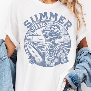 May include: White t-shirt with a blue graphic featuring a skeleton wearing a hat and sunglasses, holding a surfboard. The design includes the words "SUMMER Time" in a circular design, evoking a beach theme.